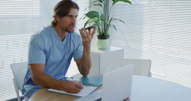 Male doctor in scrubs consulting via phone beside open laptop at office desk - Download Free Stock Images Pikwizard.com