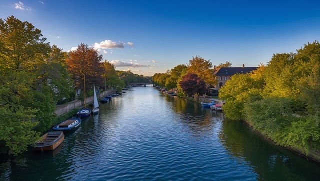 Golden hour canal in small town with moored boats, tree-lined river and bridge view