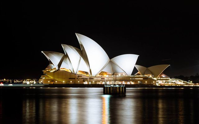 This image showcases the Sydney Opera House illuminated against the night sky, its architectural sails reflecting off the water. Useful in travel guides, architectural studies, Australian tourism promotion, and travel blogs.