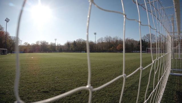 Sunlit soccer goal net framing empty grass pitch with stadium floodlights and benches