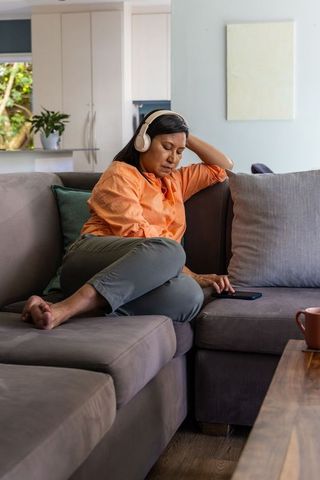 Mature woman relaxing on sofa wearing wireless headphones and checking smartphone at home