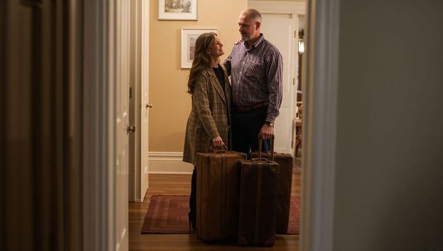 Middle-aged couple holding vintage leather suitcases in warm entryway, plaid coat and checked shirt