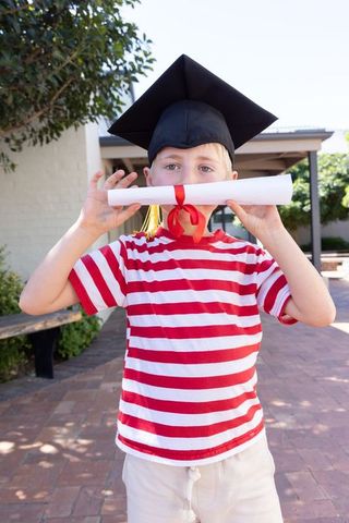 Young boy celebrating graduation wearing cap holding diploma in sunlit school courtyard