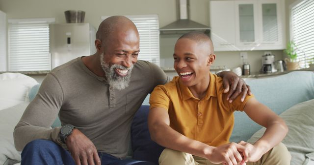 Senior man with gray beard and teenage boy laughing together while sitting on couch in modern and bright living room. Warm, joyful moment showcasing affectionate family bond, perfect for illustrating themes of family, intergenerational relationships, happiness, and home life. Ideal for advertisements, family-oriented content, blog posts on parenting or family dynamics, and health or retirement promotions.