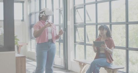 Woman wearing VR headset while coworker using tablet in bright modern coworking office