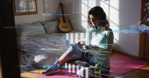 Young woman sitting on yoga mat holding phone with data overlay in sunlit bedroom