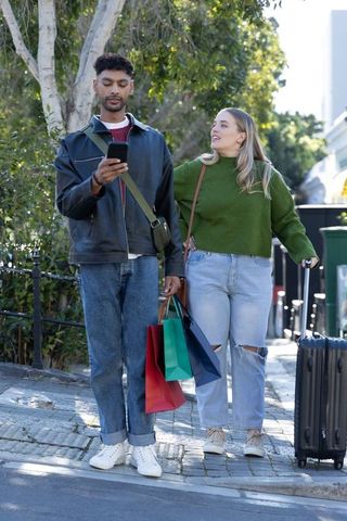 Young couple strolling in city with shopping bags and rolling suitcase, man checking phone