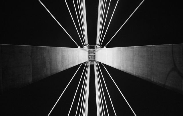Black and white shot of suspension bridge cables taken from below at night. Effective for illustrating concepts like urban architecture, engineering marvels, geometric structure, symmetry, and nighttime cityscapes. Suitable for use in architectural magazines, engineering websites, design portfolios, and urban-themed projects.