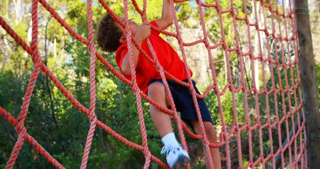 Boy actively climbing rope net in outdoor adventure park on sunny day. Ideal for use in educational materials, advertisements promoting physical fitness, playground equipment catalogs, or childcare and nature activity guides. Image conveys energy, childhood fun, and outdoor adventure.