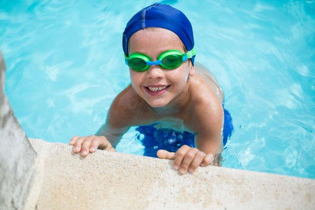 Young boy enjoying a swim in a pool, wearing green goggles and a blue swim cap, smiling at the camera. Perfect for summer activities, children's sports, swimming lessons, and healthy lifestyle promotions.