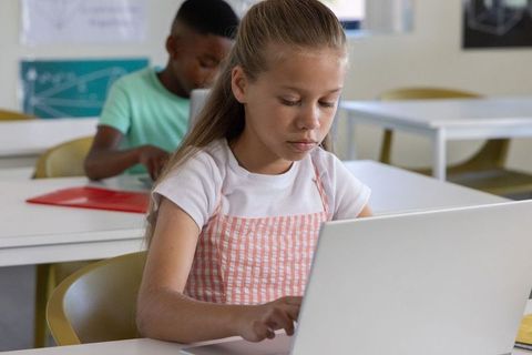 Young girl concentrating on laptop in bright classroom with classmate learning together