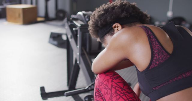 Woman weary from intense workout, resting her head on her knees in gym environment. Can be used for themes around fitness, exercise, resilience, pushing limits, and the challenge of workouts.