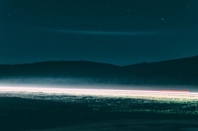 Nighttime scene capturing long exposure of light trails passing through rural landscape under starry sky. Ideal for concepts of tranquility, nature’s beauty, night photography, travel, and serenity in outdoor settings.