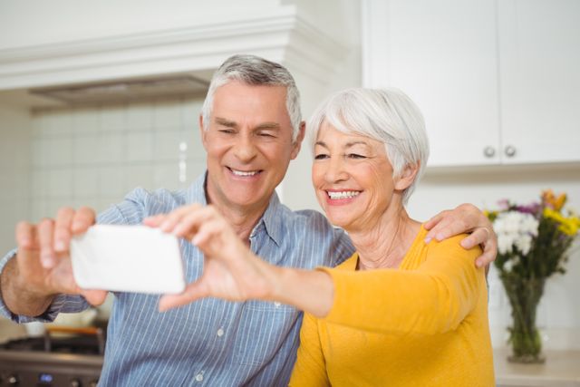 Senior couple smiling and taking a selfie in their kitchen. Ideal for use in advertisements promoting technology use among seniors, family bonding, and domestic life. Suitable for articles on aging, modern technology, and lifestyle.