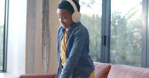 African american boy dancing on leather couch with white headphones enjoying music indoors