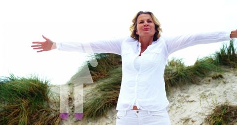 Mature woman embracing sea breeze, spreading arms on sandy dune in white outfit