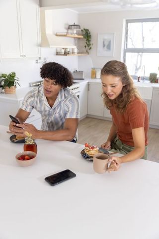 Young couple sharing pancakes and checking smartphone at sunlit modern kitchen countertop