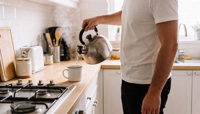 Morning man pouring steaming kettle into enamel mug on bright home kitchen counter