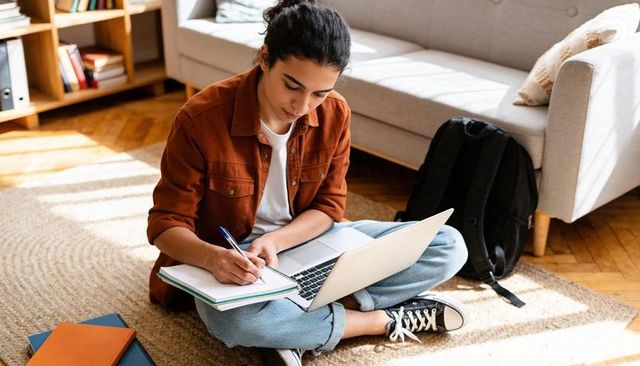 Student studying and taking notes with laptop on sunlit living room floor, backpack nearby