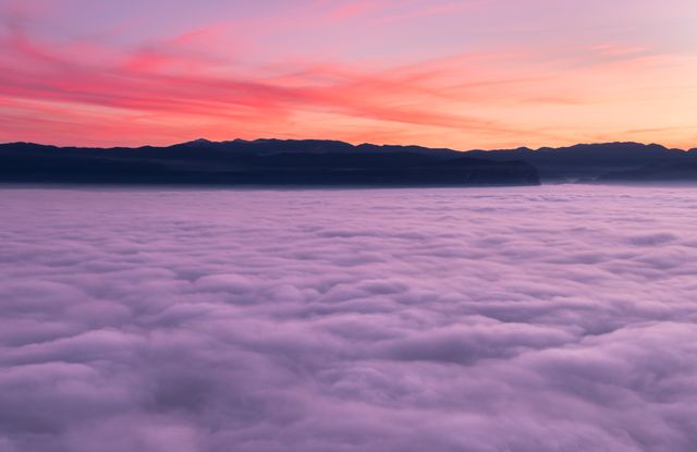 Lavender clouds cover a fog-filled valley illuminated by the gentle hues of sunrise, with dark mountain silhouettes on the horizon. Ideal for creating calming and inspirational designs, wellness and mindfulness promotions, nature and travel brochures.