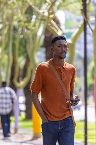 Urban african man walking tree-lined sidewalk holding smartphone and tan crossbody bag in sunlight