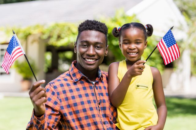 Father and daughter smiling and holding USA flags in a park. Perfect for themes related to family bonding, patriotism, Independence Day celebrations, and outdoor activities. Suitable for advertisements, social media posts, and articles about family life and national holidays.
