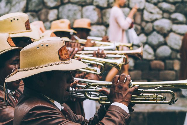 Outdoor scene shows a brass band performing in front of a stone wall. Musicians wear brown traditional cowboy hats and brown uniforms. Ideal for use in content related to cultural celebrations, parades, traditional music, and outdoor events.