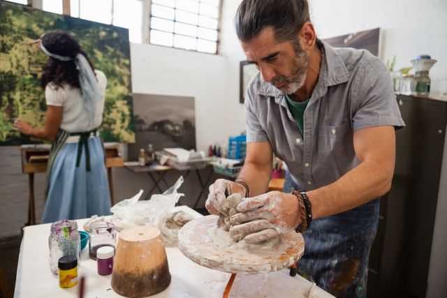 Man focused on molding clay on a pottery wheel while a woman paints on a canvas in the background. Ideal for illustrating artistic processes, creative workshops, teamwork in art, and the atmosphere of an art studio. Suitable for use in articles about pottery, painting, art classes, and creative hobbies.
