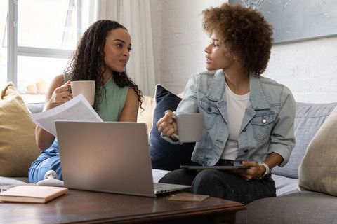 African american women collaborating on laptop and discussing documents in cozy living room