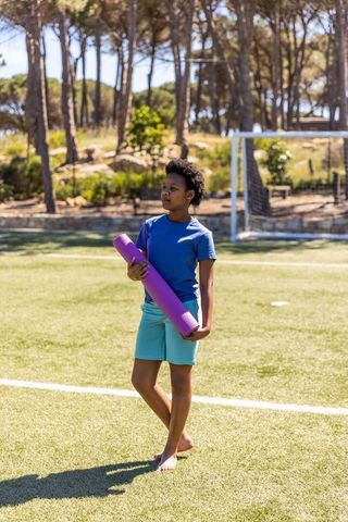 African american teen carrying purple yoga mat on sunny soccer field near pine trees