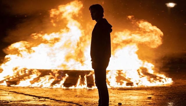 Silhouetted man standing before roaring bonfire on wet urban asphalt at night cinematic