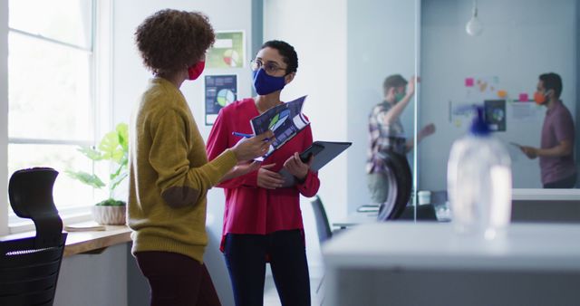 Two coworkers, a woman with curly hair holding documents and a woman with a red blouse holding a tablet, are discussing a project in a modern office. They are both wearing masks, indicating adherence to pandemic safety measures. In the background, another coworker is seen writing on a whiteboard. This image is useful for illustrating themes of teamwork, collaboration, safety measures during a pandemic, and a modern business environment.