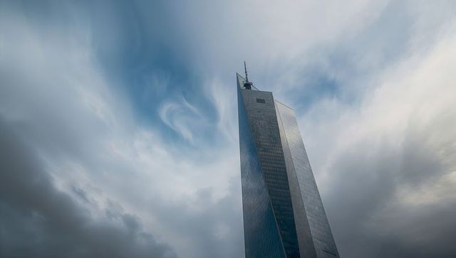 Glass skyscraper rising into dramatic cloudy sky reflecting city skyline with sharp spire