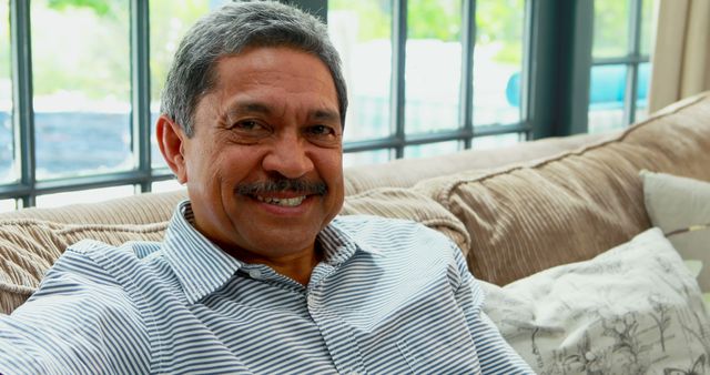 Senior man in striped shirt is relaxing on a couch with a smile on his face. The setting appears to be a cozy home with large windows allowing natural light inside. This image can be used for themes related to senior living, happiness, relaxation, home comfort, retirement lifestyle, or family life.