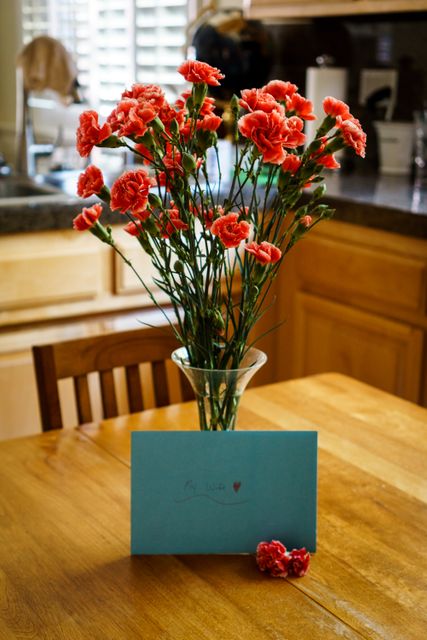 Red carnations in glass vase placed on a wooden dining table in bright kitchen. Beside the vase, a blue greeting card with handwriting is visible, suggesting a personal touch and special occasion. Suitable for themes related to love, celebrations, home decor, and family gatherings.