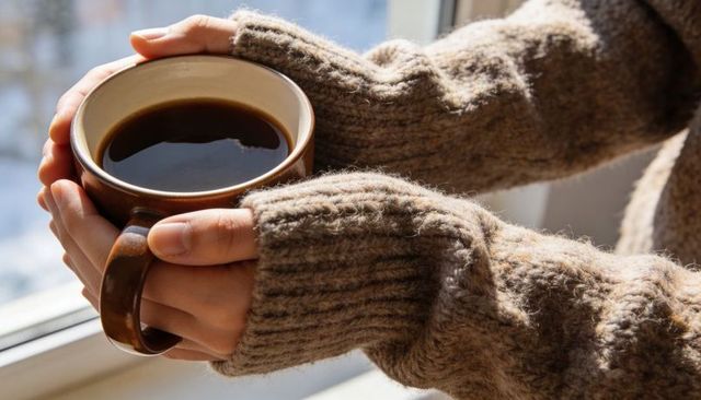 Cradling hands warming on clay mug of black coffee in chunky knit sleeves by sunlit window