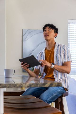 Asian man sitting at kitchen island using tablet tapping screen, modern home lifestyle