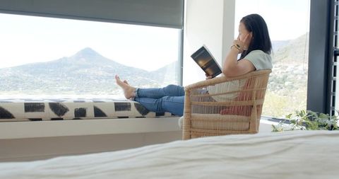 Asian woman reading by panoramic window with mountain view in cozy rattan chair minimal