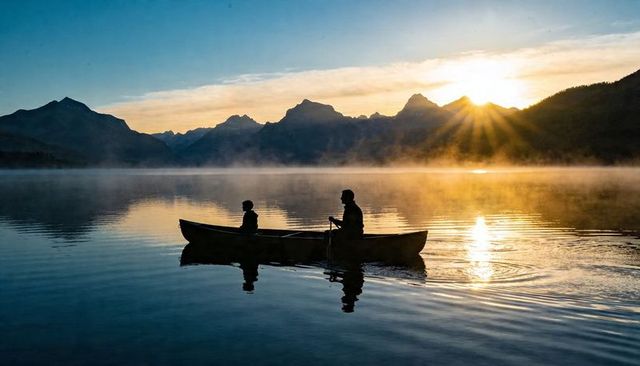 Paddling canoe at sunrise with adult and child silhouette on misty mountain lake