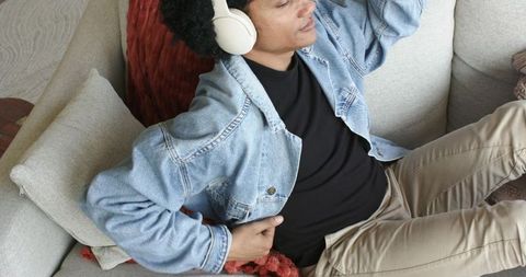 Mid-adult man relaxing on grey sofa with headphones and denim jacket, cozy living room vibe