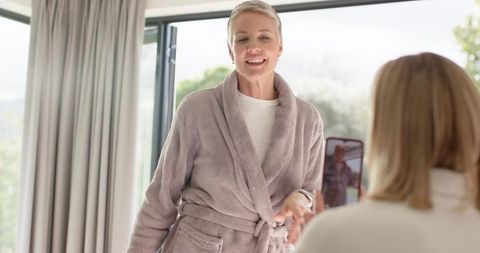 Mature woman posing in plush robe while friend filming on smartphone in sunlit home