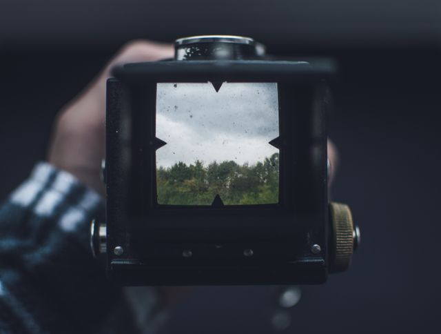Close-up of hands holding vintage camera while looking through viewfinder. Viewfinder shows a scenic landscape with trees and a cloudy sky. Ideal for themes of nostalgia, retro photography, nature, outdoor activities, and hobbyist photography.
