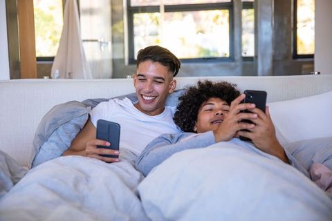 Diverse male couple relaxing in bed with smartphones
