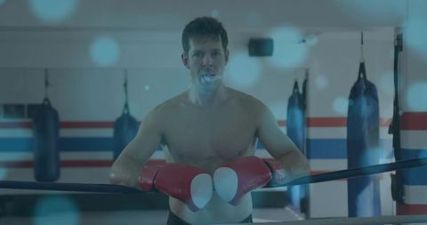 Leaning shirtless boxer on ring ropes wearing red gloves and mouthguard