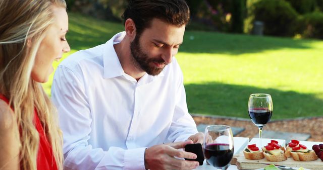A young Caucasian couple enjoys a romantic outdoor picnic with wine and appetizers, with copy space. Their relaxed demeanor and smiles suggest a moment of leisure and connection in a serene setting.