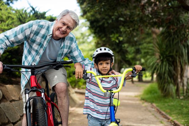 Grandfather and grandson standing with their bicycles in a park, enjoying a sunny day. The grandfather is smiling, and the grandson is wearing a helmet, showcasing a moment of family bonding and outdoor activity. Ideal for use in family-oriented advertisements, health and wellness campaigns, or articles about active lifestyles and intergenerational relationships.