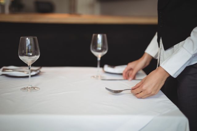 Image depicting a waitress setting an elegant table in a restaurant. The scene includes wine glasses, a white tablecloth, and neatly arranged silverware. Suitable for use in hospitality industry promotions, restaurant advertising, service training materials, and dining experience blogs.