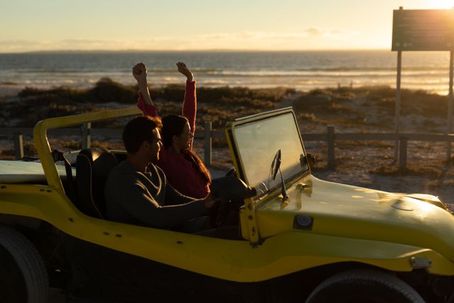Happy caucasian couple driving beach buggy by sea at sunset, woman raising arms. beach stop off on romantic summer holiday road trip.