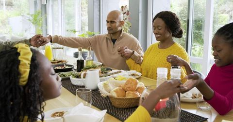 Multiethnic family holding hands bowing heads at sunlit dining table giving thanks
