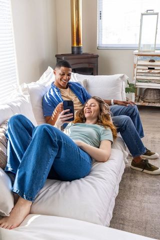 Young couple relaxing on white sofa sharing smartphone in sunlit cozy modern living room